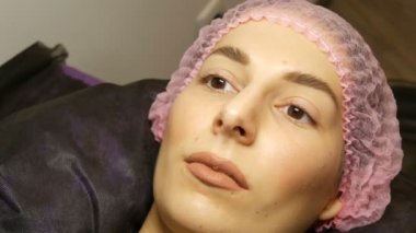 Geniş kıllı kaşları, kahverengi gözleri ve pembe şapkasıyla dudaklarında mat bej ruj izi olan güzel bir kadının yüzü güzellik salonunda kirpikleri kaplama işleminden önce.