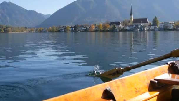 A peaceful picture a wooden boat with an oar floats on beautiful mountain lake Tegernsee against backdrop of Alpine mountains and the picturesque container of the church. Ferryman ferries people — Stock Video