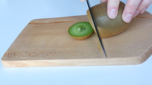 Frauenhände schneiden reife Kiwi auf einem Küchenholzbrett mit schwarzem Keramikmesser auf einem weißen Tisch — Stockvideo