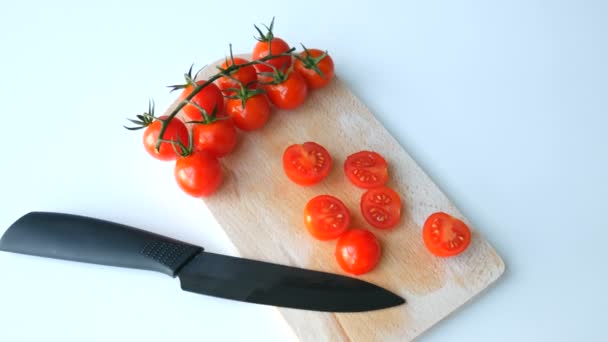 Elegante cuchillo de cocina de cerámica y pequeños tomates cherry rojos maduros en racimos y parcialmente cortados en el tablero de la cocina — Vídeo de stock