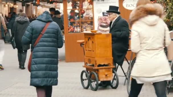 Norymberga, Niemcy - 10 grudnia 2019: Starszy męski szlifierka organów w czarnym płaszczu i kapeluszu gra na starym drewnianym narządzie lufy na pustkowiu miasta. Spacerujący tłum ofiarowuje monety i pieniądze — Wideo stockowe