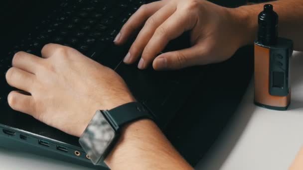 Un hombre trabaja en el portátil. Mens manos con un reloj inteligente y un cigarrillo electrónico están escribiendo en el teclado . — Vídeo de stock