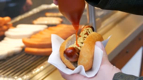 Hot dog. Il cibo di strada non è salutare. La mano maschile tiene una salsiccia in panino e la versa con ketchup in appositi contenitori — Video Stock