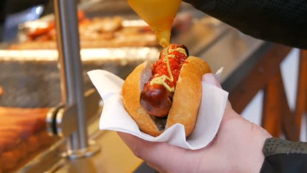 Hot Dog. Comida chatarra callejera. La mano masculina sostiene una salchicha en bollo y la vierte con mostaza en recipientes especiales — Vídeo de stock