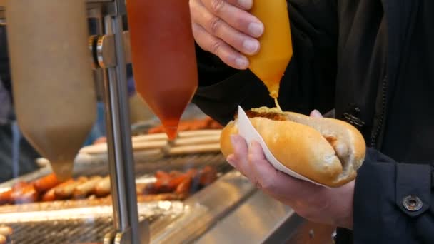 Hot Dog. Comida de rua. A mão masculina segura uma salsicha em pão e derrama-a com mostarda em recipientes especiais. — Vídeo de Stock