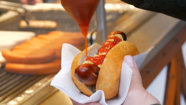 Hot Dog. La comida callejera no es saludable. La mano masculina sostiene una salchicha en bollo y la vierte con ketchup en recipientes especiales — Vídeos de Stock