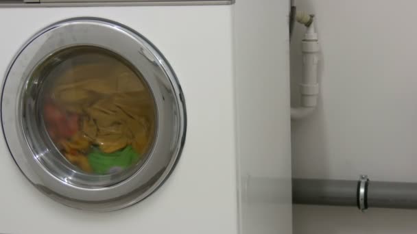 La ropa de varios colores se lava en una lavadora blanca en la sala de lavandería . — Vídeo de stock