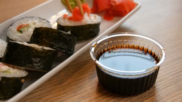 Grandes rollos de sushi con salmón en nori, palitos de cangrejo, pepino, queso Filadelfia sumergido en un tazón de salsa de soja con la ayuda de palos de bambú de cerca ver — Vídeos de Stock