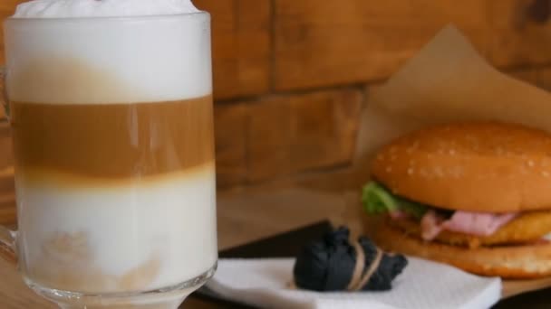 Delicioso café con leche recién hecho en una mesa en un café. Espuma de leche de café con leche en vidrio especial largo transparente. En el fondo es una deliciosa hamburguesa con lechuga, chuleta de carne. Comida rápida poco saludable — Vídeo de stock