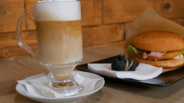 Delicioso café con leche recién hecho en una mesa en un café. Espuma de leche de café con leche en vidrio especial largo transparente. En el fondo es una deliciosa hamburguesa con lechuga, chuleta de carne. Comida rápida poco saludable — Vídeos de Stock