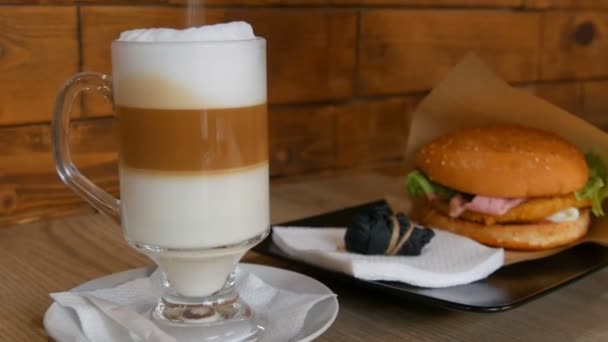 Delicioso café con leche recién hecho en una mesa en un café. Espuma de leche de café con leche en vidrio especial largo transparente. El azúcar espolvorea en la bebida. En el fondo es deliciosa hamburguesa con lechuga, chuleta de carne — Vídeo de stock