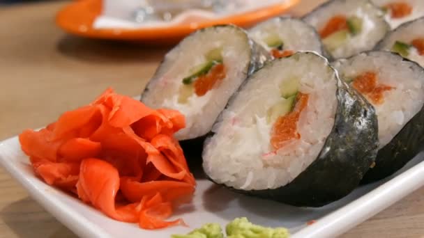 Grandes rollos de sushi en un nori con salmón, palitos de cangrejo, pepinos, queso filipino. Muy cerca está el wasabi verde y el jengibre rojo. Comida japonesa — Vídeo de stock