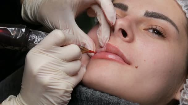 Microblading τατουάζ χειλιών με ένα ειδικό χρωματισμό κόκκινη χρωστική ουσία που διορθώνει το χρώμα των χειλιών σε μια κλινική κοσμετολογίας. Μόνιμη διαδικασία μακιγιάζ χείλη εφαρμογή χρωστική μακιγιάζ στα χείλη με ένα μηχάνημα τατουάζ — Αρχείο Βίντεο