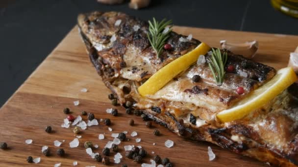 Delicioso pescado fresco, caballa ahumada en una tabla de cortar de madera junto a los tomates cherry, ajo, sal gruesa y pimienta, decorado con rodajas de limón y ramitas de romero verde — Vídeos de Stock