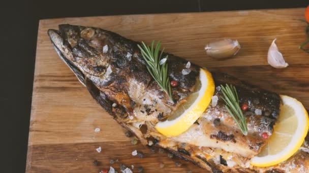 Delicioso pescado fresco, caballa ahumada en una tabla de cortar de madera junto a los tomates cherry, ajo, sal gruesa y pimienta, decorado con rodajas de limón y ramitas de romero verde — Vídeos de Stock