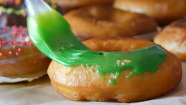 Una fila de rosquillas redondas más grandes recién fritas en una mesa de cocina casera. Un cepillo especial de silicona de cocina aplica un esmalte verde brillante a la superficie de la dona. Grasa, comida chatarra, comida rápida de cerca — Vídeos de Stock