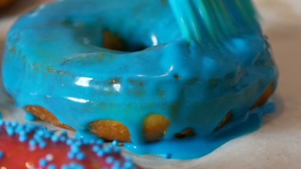 Een rij grotere ronde vers gebakken donuts op een zelfgemaakte kooktafel. Een speciale keuken siliconen borstel brengt een helder blauw glazuur aan op het oppervlak van de donut. Vet, junk food, fast food close-up — Stockvideo