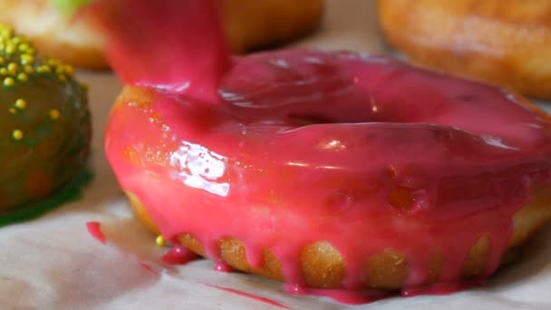 Eine Reihe größerer runder, frisch gebratener Donuts auf einem heimischen Kochtisch. Eine spezielle Silikonbürste für die Küche trägt eine leuchtend rosa Glasur auf die Oberfläche des Donuts auf. Fettiges, Junk Food, Fast Food — Stockvideo
