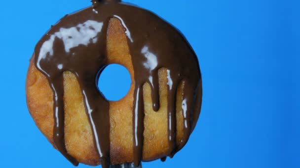 Grande donut redondo em um garfo no qual o esmalte de cobertura de chocolate flui em um contexto azul — Vídeo de Stock