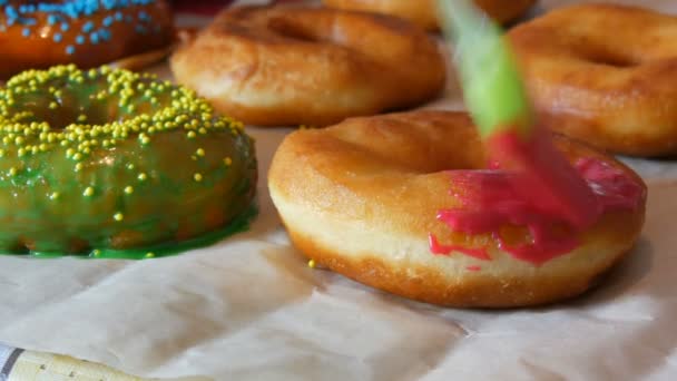 A row of larger round freshly fried donuts on a home cooking table. A special kitchen silicone brush applies a bright pink glaze to the surface of the donut. Fatty, junk food, fast food — Stock Video