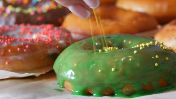 Large multi-colored fresh fried donuts in a row on a table. A beautiful donut with green icing is sprinkled with a special colored powder for decoration of sweets — Stock Video
