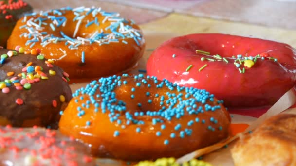 Grands beignets frits frais multicolores en rangée sur une table. Un beau beignet avec glaçage rose est saupoudré d'une poudre colorée spéciale pour la décoration de bonbons — Video