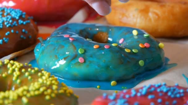 Große bunte, frisch gebratene Donuts in einer Reihe auf einem Tisch. Ein schöner Donut mit blauem Zuckerguss wird mit einem speziellen farbigen Puder zur Dekoration von Süßigkeiten bestreut — Stockvideo