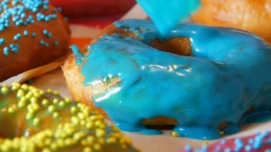 Bir sıra büyük yuvarlak taze kızarmış çörekler bir ev yemeği masasında. Özel bir mutfak silikon fırçası donutun yüzeyine parlak mavi bir jöle sürer. Yağlı, abur cubur, fast food. Yaklaş.