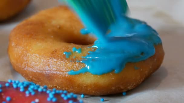 Uma fileira de rosquinhas fritas redondas maiores em uma mesa de culinária caseira. Uma escova de silicone de cozinha especial aplica um esmalte azul brilhante à superfície do donut. Gordo, junk food, fast food close-up — Vídeo de Stock