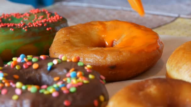 Een rij grotere ronde vers gebakken donuts op een zelfgemaakte kooktafel. Een speciale keuken siliconen borstel brengt een fel oranje glazuur aan op het oppervlak van de donut. Vet, junk food, fast food close-up — Stockvideo