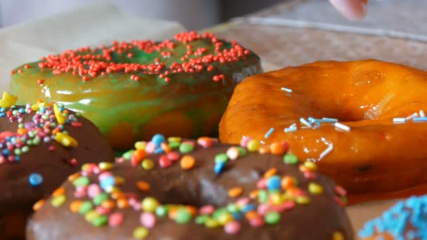 Große bunte, frisch gebratene Donuts in einer Reihe auf einem Tisch. Ein schöner Donut mit orangefarbener Zuckerglasur wird mit einem speziellen farbigen Puder bestreut, um Süßigkeiten zu dekorieren — Stockvideo