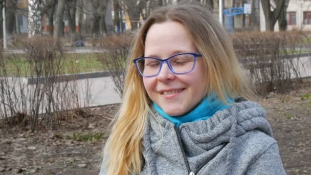 Красивая молодая женщина с светлыми волосами и голубыми глазами в очках сидит в весеннем парке и улыбается — стоковое видео