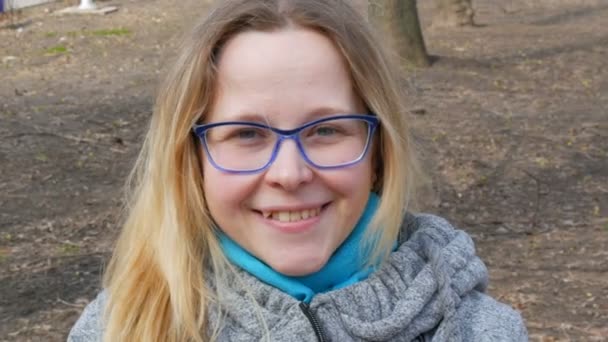 Mooie jonge vrouw met blond haar en blauwe ogen in bril zit in een lentepark en glimlacht — Stockvideo