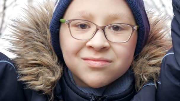Милый веселый мальчик-подросток 10 лет в очках играет и катается на качелях на детской площадке ранней весной — стоковое видео