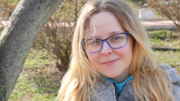 Hermosa joven con el pelo rubio y ojos azules en gafas se sienta en un parque de primavera y sonríe — Vídeos de Stock