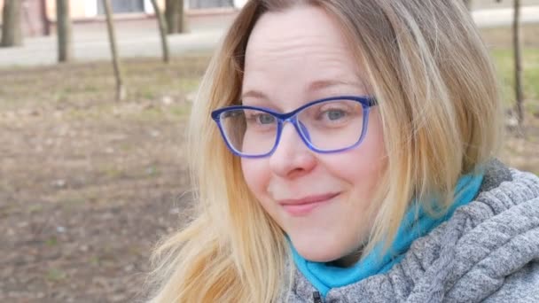 Retrato de una hermosa joven con el pelo rubio y los ojos azules con gafas sentadas en un parque de primavera y sonriendo y haciendo caras — Vídeos de Stock