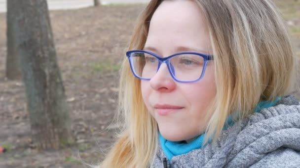 Красивая молодая женщина с светлыми волосами и голубыми глазами в очках сидит в весеннем парке и улыбается — стоковое видео