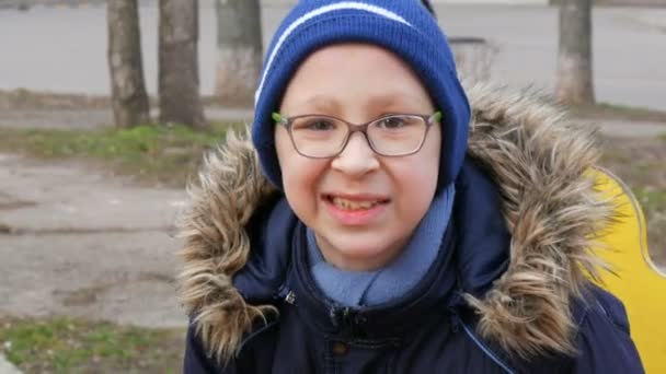 Милий веселий хлопчик підліток десяти років в окулярах грає і їде на гойдалці на дитячому майданчику ранньої весни — стокове відео