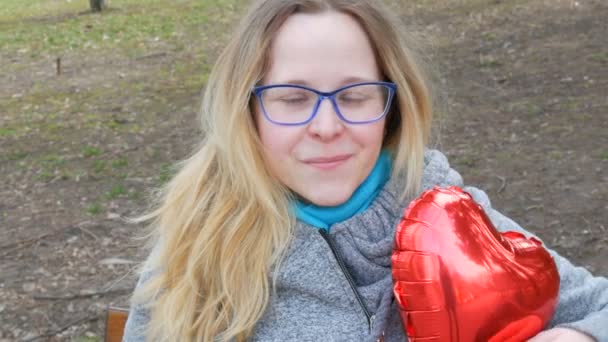 Retrato de una hermosa joven con el pelo rubio y los ojos azules con gafas sentadas en un parque de primavera y sonriendo. Globo rojo en forma de corazón en las manos — Vídeos de Stock