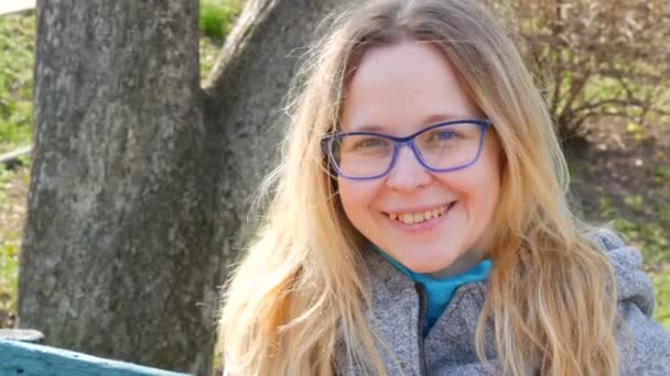 Sarı saçlı ve gözlüklü mavi gözlü güzel bir genç kadın bahar parkında oturuyor ve gülümsüyor. — Stok video