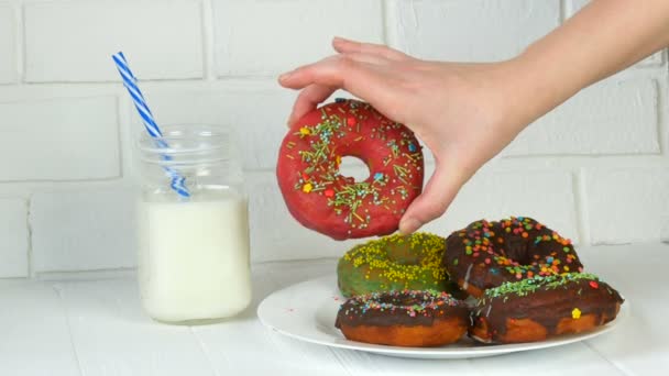Женские руки берут большой американский пончик с разноцветным порошком на фоне белой кирпичной стены и молока в банке. Нездоровая пища, диабет — стоковое видео