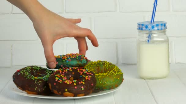 Female hands take a big American donut with multi-colored powder on a background of a white brick wall and milk in a jar. Junk food, diabetes — Stock Video