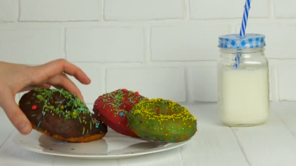 Female hands take a big American donut with multi-colored powder on a background of a white brick wall and milk in a jar. Junk food, diabetes — Stock Video