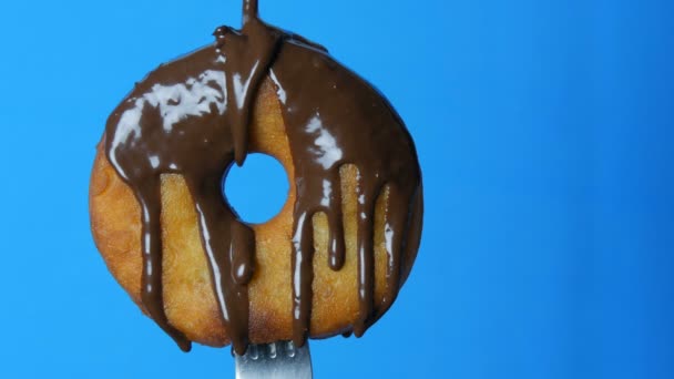 Donut redondo grande en un tenedor en el que el glaseado de chocolate y polvo azul giran sobre un fondo azul — Vídeos de Stock