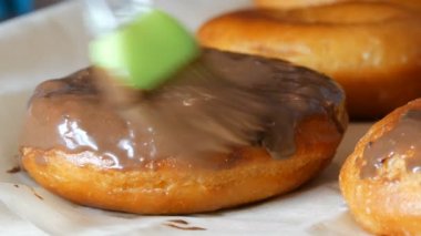 Bir sıra büyük yuvarlak taze kızarmış çörekler bir ev yemeği masasında. Özel bir mutfak silikon fırçası donutun yüzeyine parlak çikolata jölesi sürer. Yağlı, abur cubur, fast food. Yaklaş.