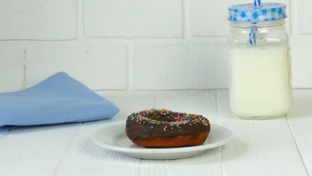 Большой шоколадный пончик с цветным порошком на белой тарелке напротив кирпичной стены рядом с банкой молока и синей салфеткой. Нездоровая пища, диабет — стоковое видео