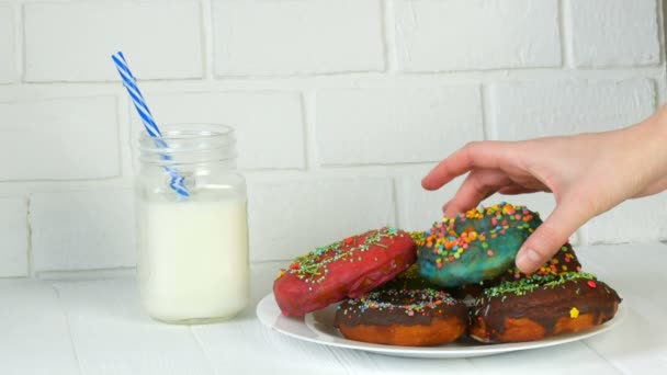 Female hands take a big American donut with multi-colored powder on a background of a white brick wall and milk in a jar. Junk food, diabetes — Stock Video