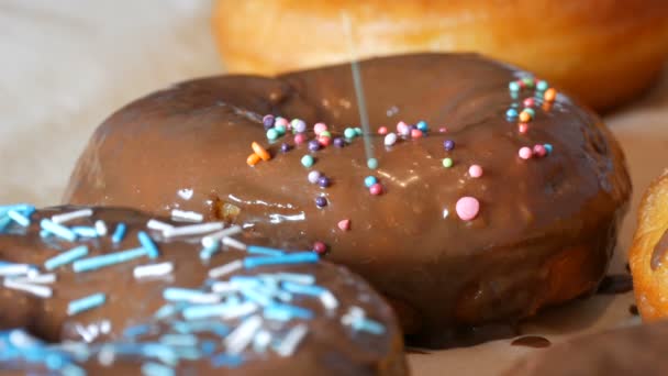 Os grandes donuts fritos frescos multicores sucessivamente em uma mesa. Um belo donut com cobertura de chocolate é polvilhado com um pó colorido especial para decoração de doces. — Vídeo de Stock