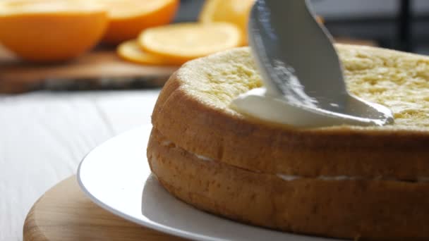 Mujer esparciendo una crema en la masa horneada. La mano femenina vierte crema blanca con una cuchara en el pastel y comienza a esparcirse en una galleta naranja o masa de bizcocho en la cocina. — Vídeos de Stock