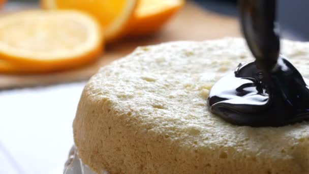 Vista de cerca. Mujer que extiende una crema de chocolate espesa o glaseado en la masa horneada con una cuchara en el pastel y comienzan a extenderse en una galleta de naranja o masa de pastel de esponja en la cocina — Vídeos de Stock